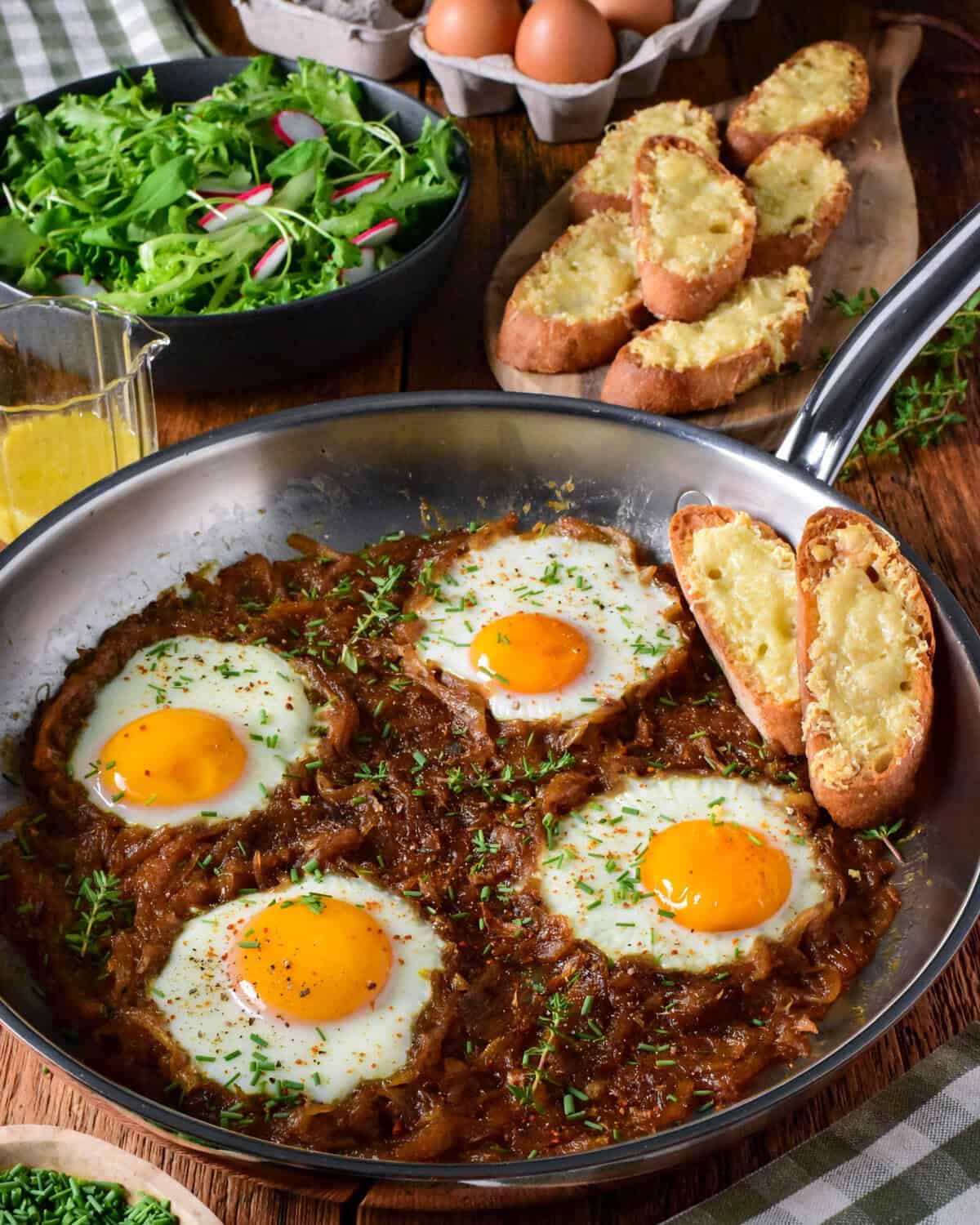 Skillet with baked eggs in caramelized onion and Gruyère cheese, served with toasted bread and fresh salad on a rustic wooden table.