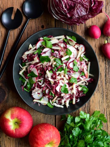 Fresh Honeycrisp apple and radicchio salad with radishes and herbs in a black bowl, on a wooden table with apples and greens around.