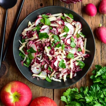 Fresh Honeycrisp apple and radicchio salad with radishes and herbs in a black bowl, on a wooden table with apples and greens around.