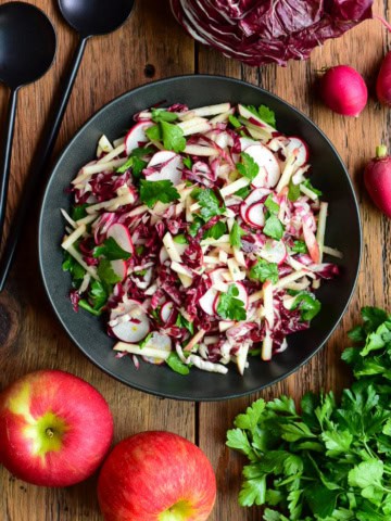Fresh Honeycrisp apple and radicchio salad with radishes and herbs in a black bowl, on a wooden table with apples and greens around.