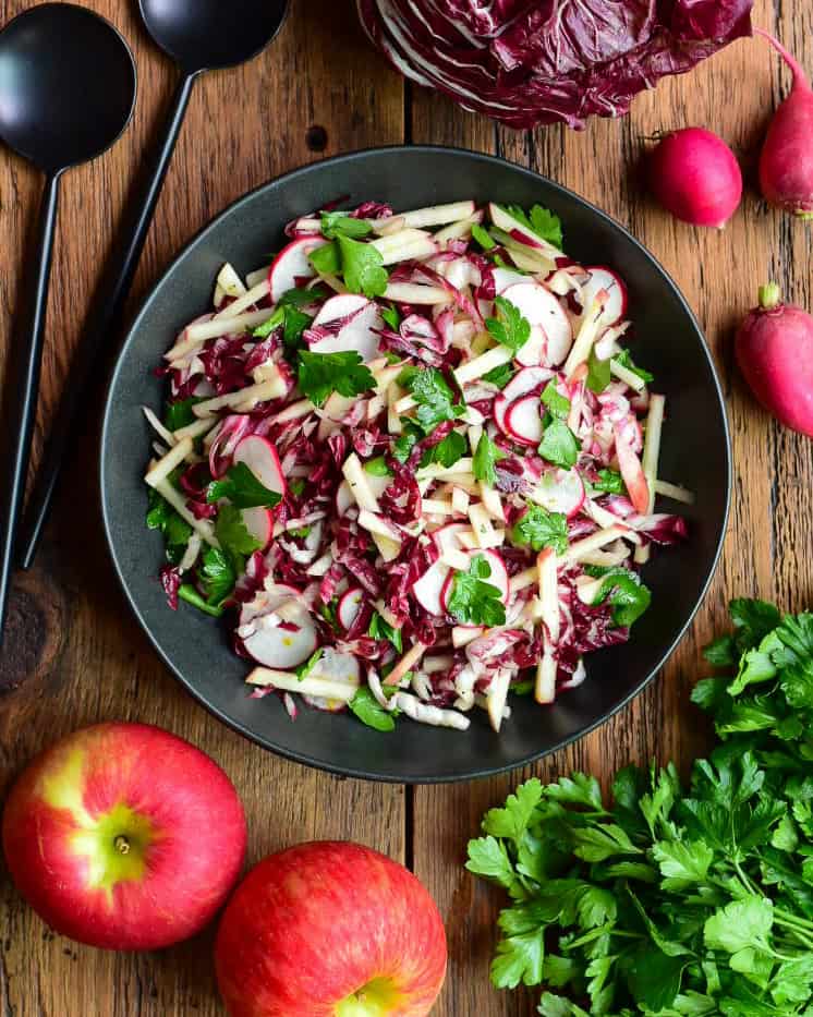 Fresh Honeycrisp apple and radicchio salad with radishes and herbs in a black bowl, on a wooden table with apples and greens around.