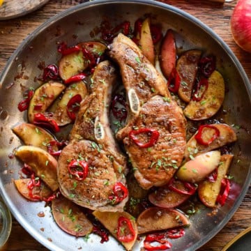 Delicious pan-seared Berkshire pork chops served with sautéed apples and red chili slices, garnished with herbs, on a rustic wooden table.