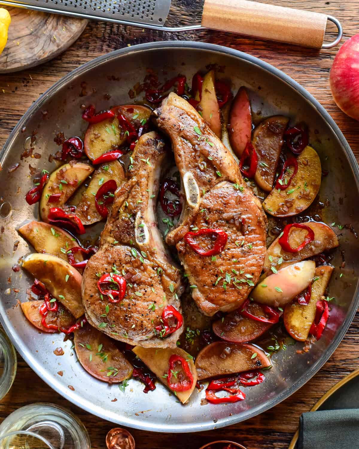Delicious pan-seared Berkshire pork chops served with sautéed apples and red chili slices, garnished with herbs, on a rustic wooden table.