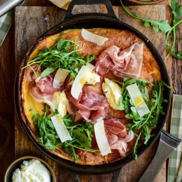 Dutch baby skillet topped with prosciutto, arugula, parmesan shavings, and creamy ricotta in a cast iron pan.