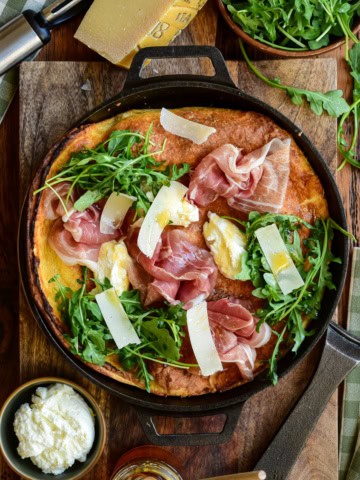 Dutch baby skillet topped with prosciutto, arugula, parmesan shavings, and creamy ricotta in a cast iron pan.
