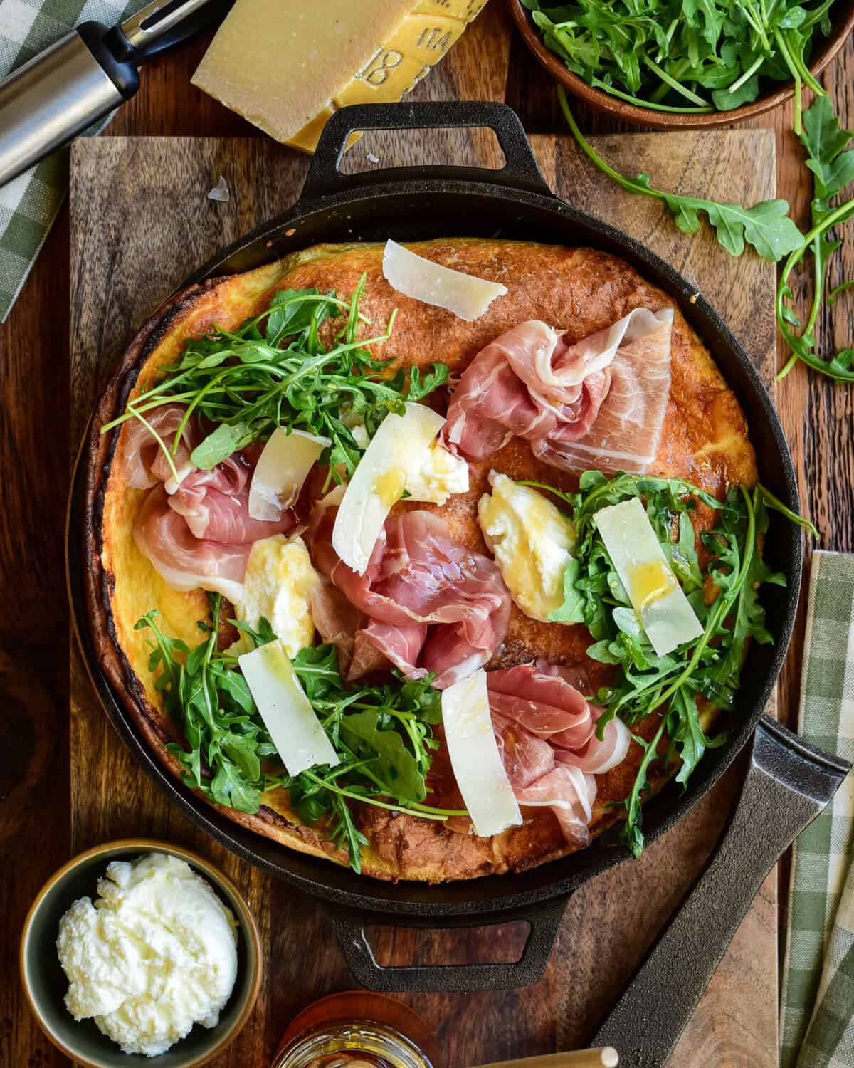 Dutch baby skillet topped with prosciutto, arugula, parmesan shavings, and creamy ricotta in a cast iron pan.