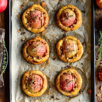 Delicious apple and cheddar puff pastry tarts baked to golden perfection, garnished with herbs, on a baking sheet with fresh apples and herbs around.