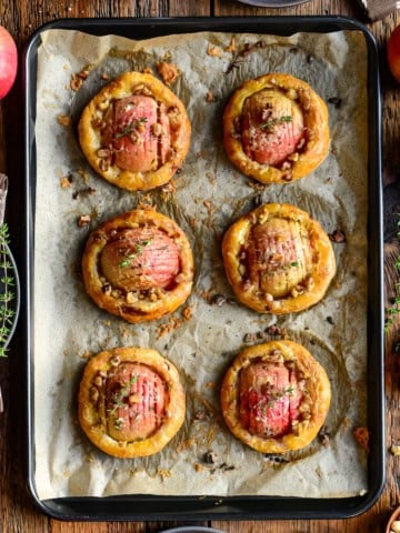 Delicious apple and cheddar puff pastry tarts baked to golden perfection, garnished with herbs, on a baking sheet with fresh apples and herbs around.