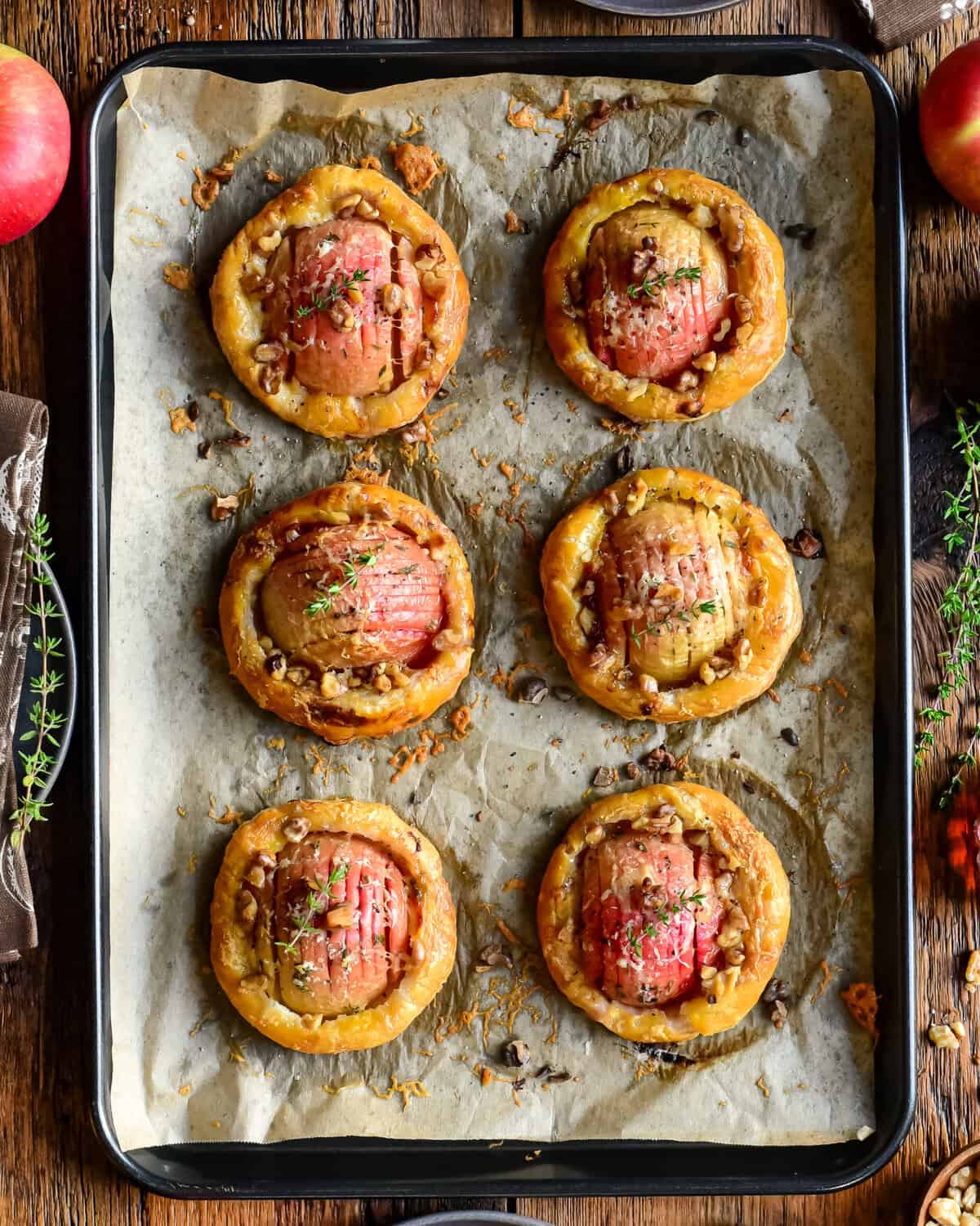 Delicious apple and cheddar puff pastry tarts baked to golden perfection, garnished with herbs, on a baking sheet with fresh apples and herbs around.
