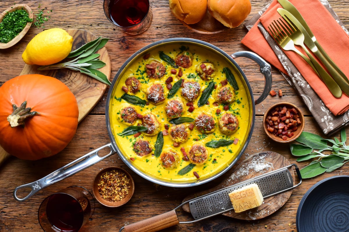 Turkey meatballs cooked in a pumpkin-sage sauce in a skillet, garnished with sage leaves and grated cheese, with pumpkins and spices in the background.
