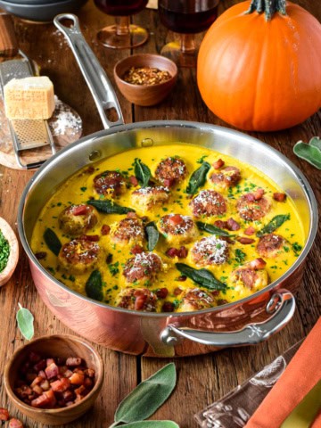 Turkey meatballs cooked in a pumpkin-sage sauce in a copper skillet, garnished with sage leaves and grated cheese, with pumpkins and spices in the background.