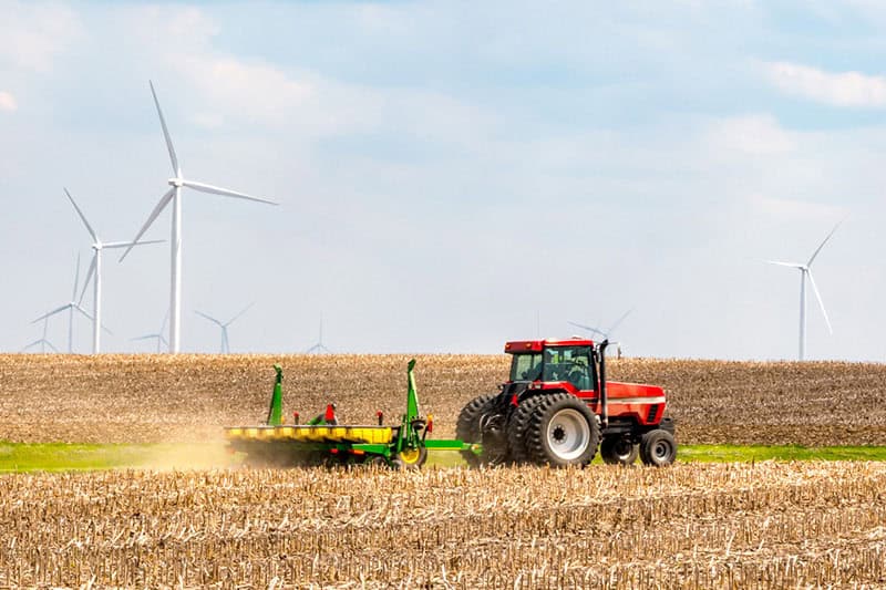 Farmer plowing land next to wind farm with wind turbines rotating in background