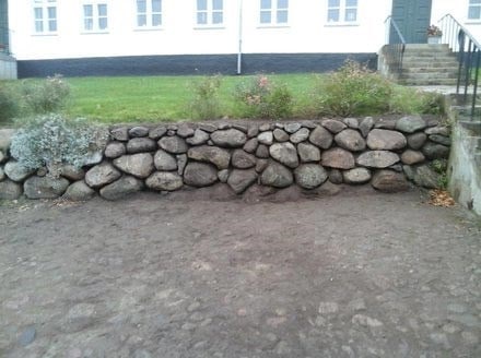 Stenmur med jord og planter foran en boligbygning.