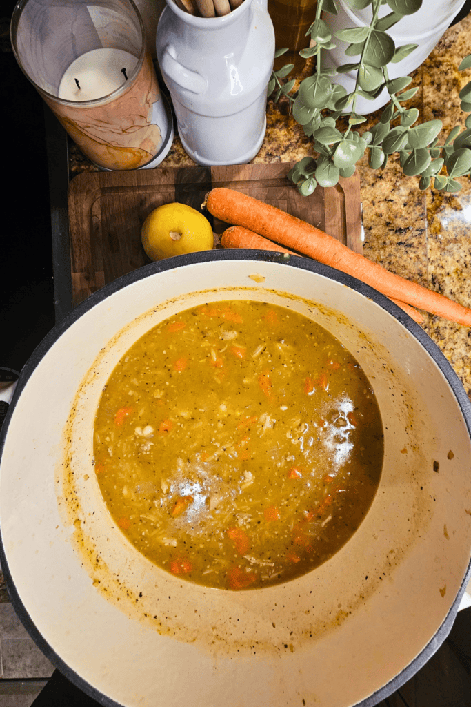 Fresh homemade lemon chicken orzo soup in a white bowl with carrots, lemon, and herbs on a kitchen countertop.