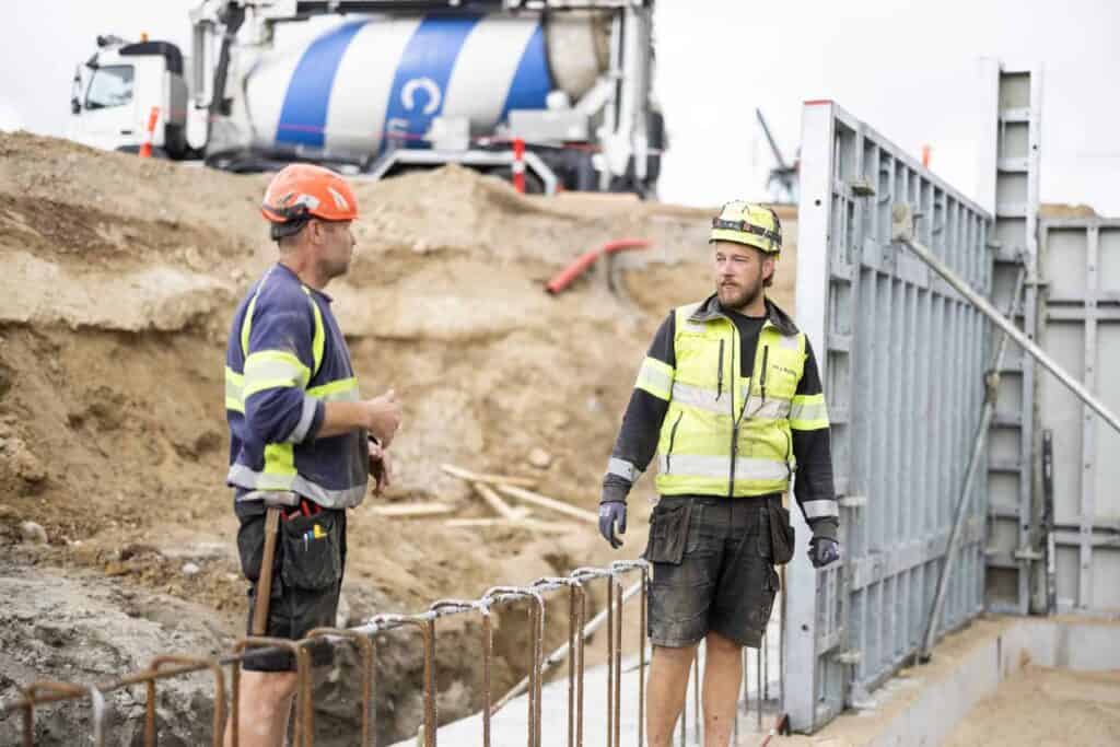 To bygningsarbejdere i sikkerhedsudstyr på byggeplads med betonblander i baggrunden.