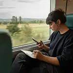 Une jeune femme assise dans un train lit une offre d’emploi sur son téléphone et prend des notes dans un petit carnet.
