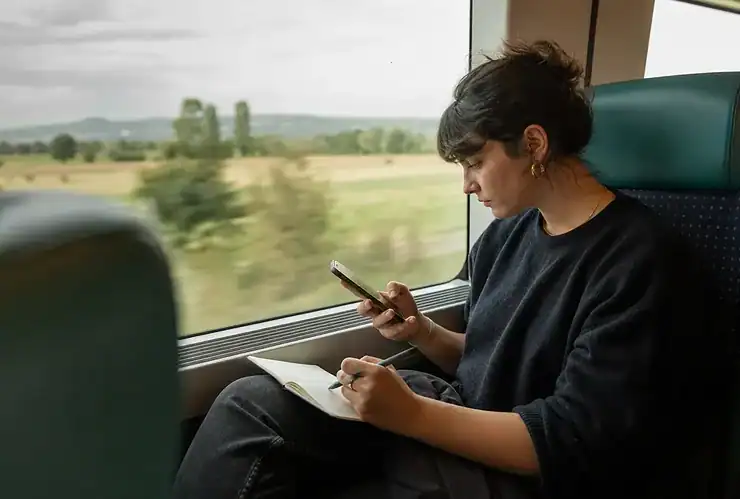 Une jeune femme assise dans un train lit une offre d’emploi sur son téléphone et prend des notes dans un petit carnet.