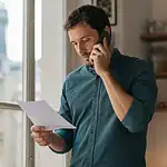 Un homme au téléphone près d’une fenêtre, une liste annotée à la main.