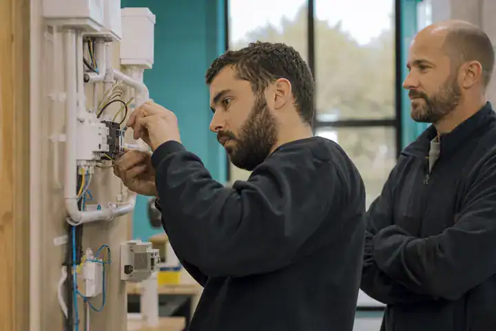 Un adulte s’exerce à une installation électrique sur un panneau de formation, sous le regard d’un formateur