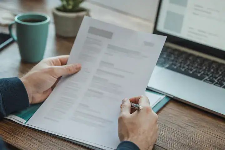 Mains relisant un CV imprimé, stylo en main, à côté d’un ordinateur portable