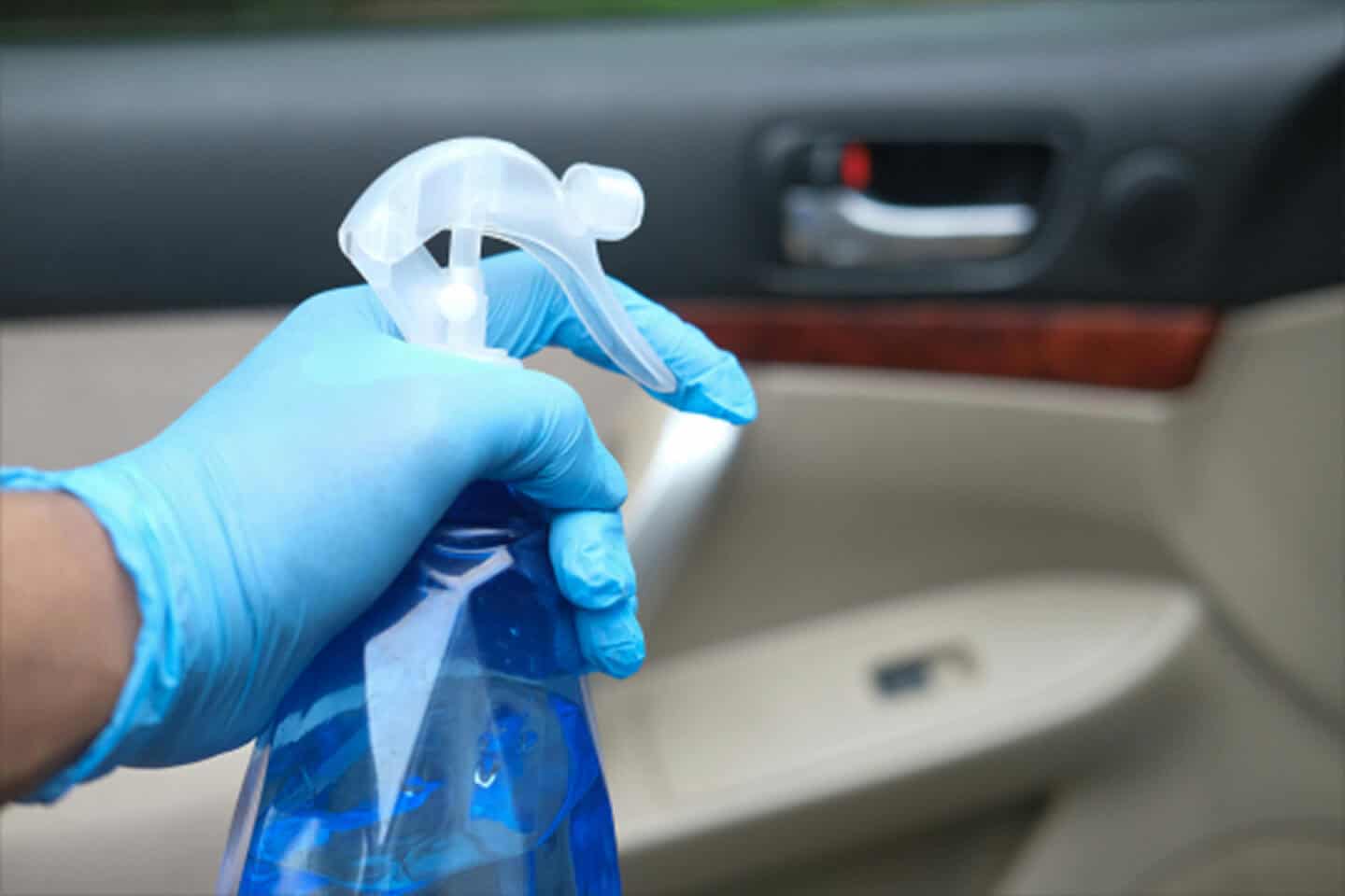 Close-up of a hand wearing a blue nitrile glove holding a blue spray bottle, ready to clean the interior door panel of a car.