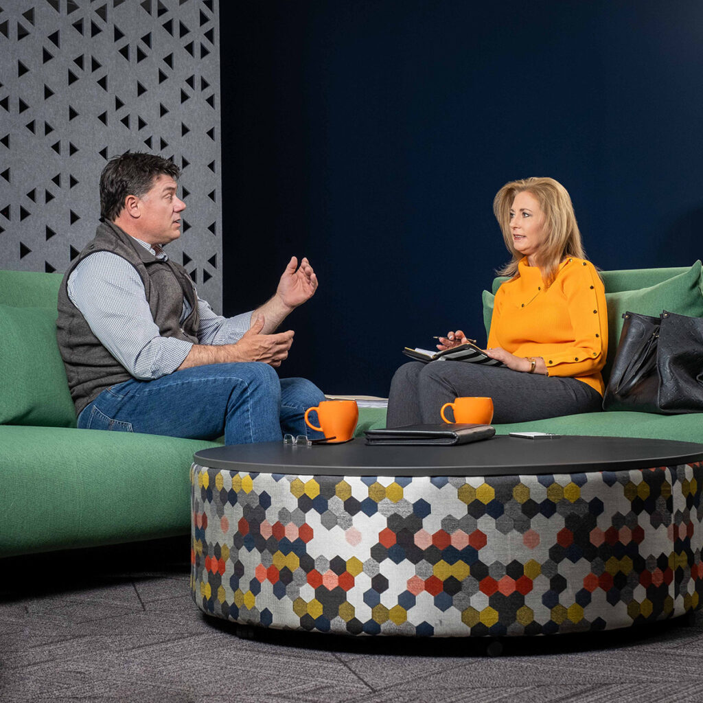 Diane consults with a coaching client while sitting on green coaches in a meeting room.