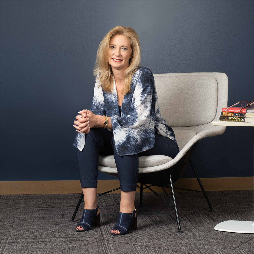 Diane Welhouse is seating on a modern style white chair, dressed in business casual clothing, smiling at the camera.