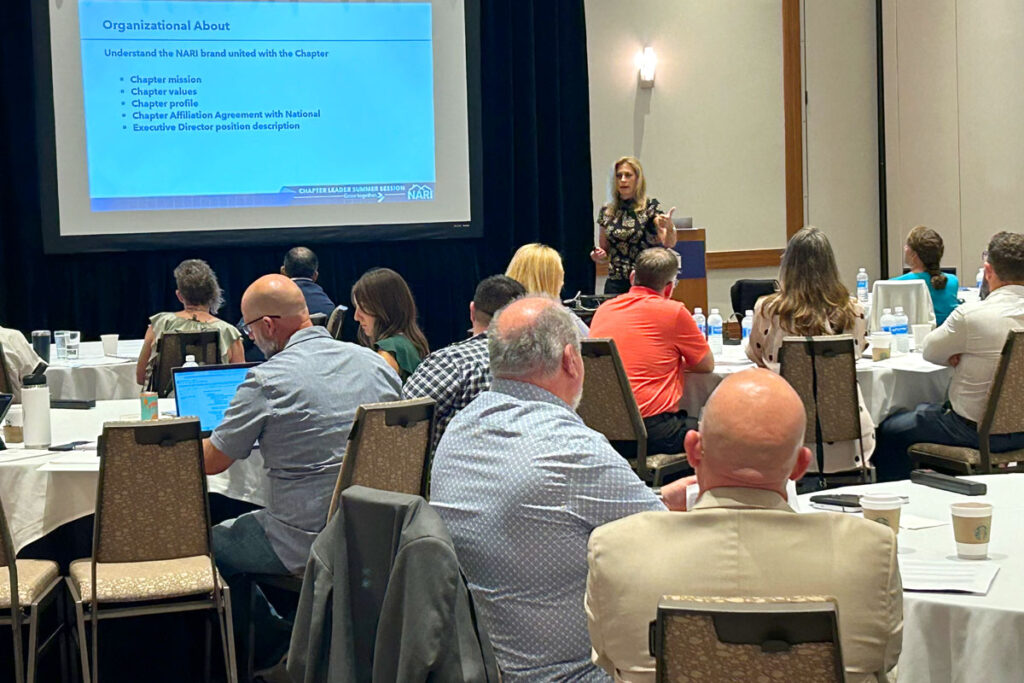 Diane Welhouse speaking in front of a crowd of business professionals, with a projected presentation on a screen beside her.