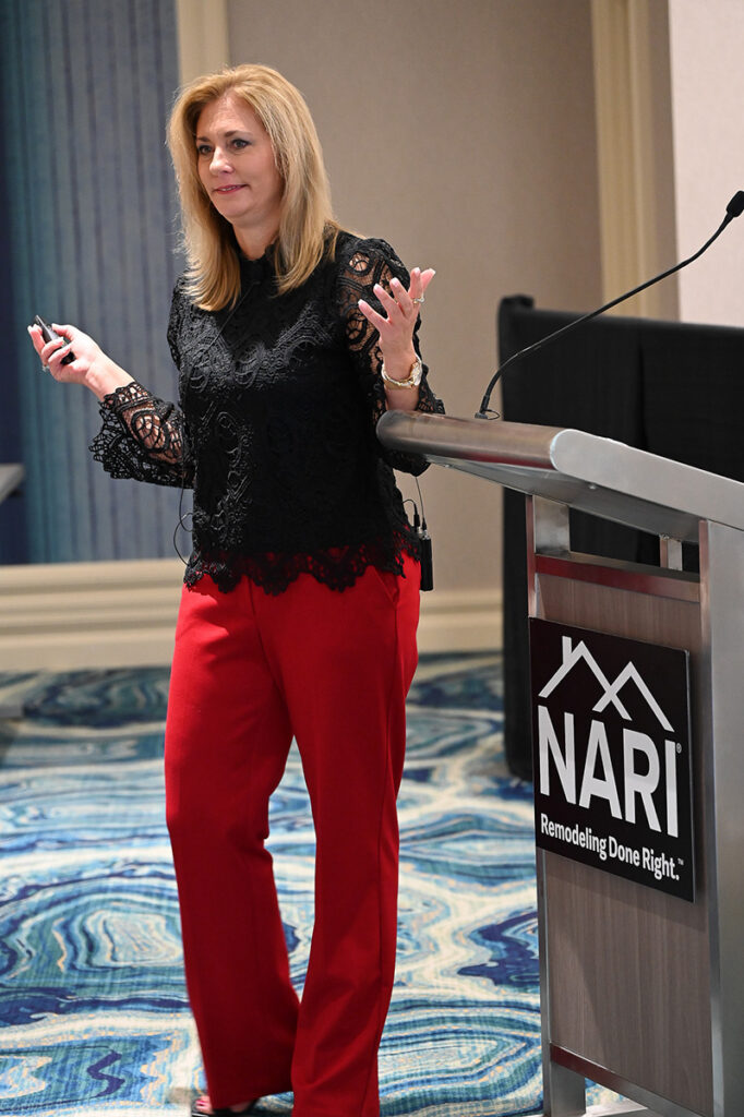 Diane Welhouse gestures during a speech, standing next to a podium, wearing a black top and red pants.
