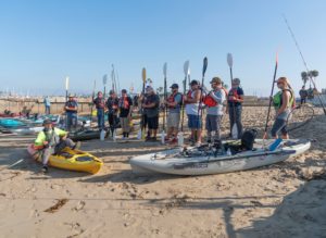 A large group of paddlers stand with their paddles vertical while a guide explains kayaking techniques