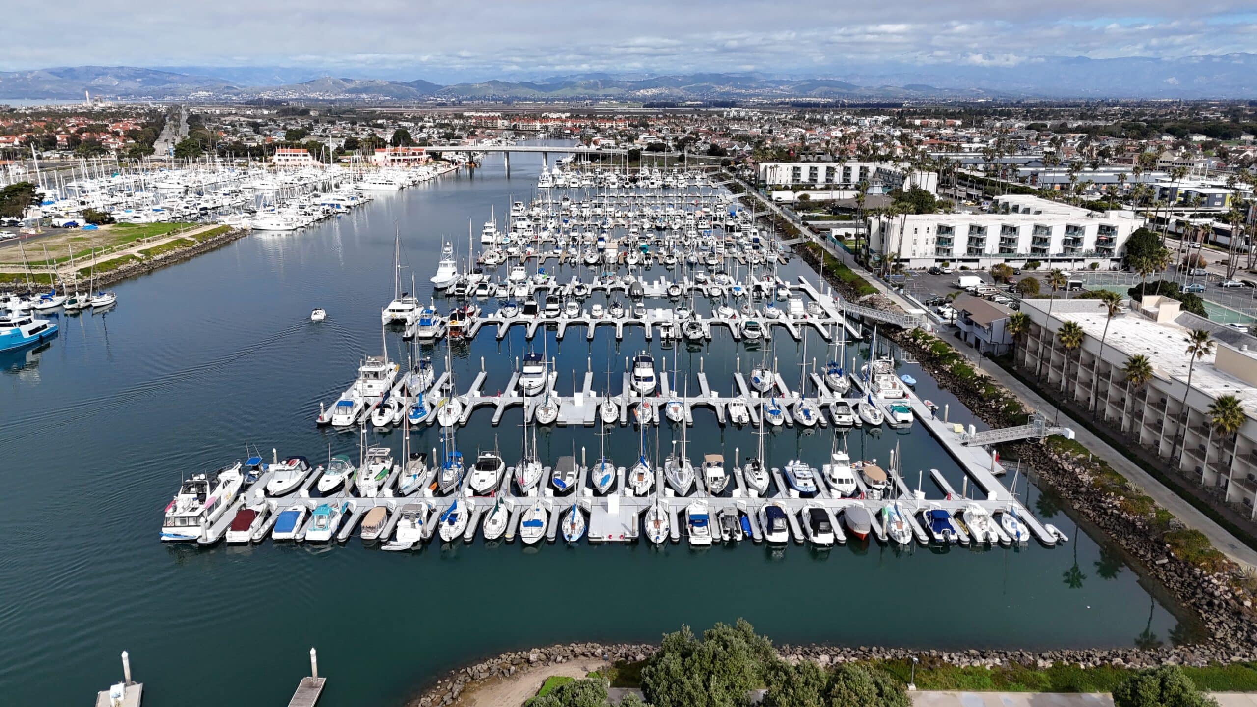 Aerial view of a marina with numerous boats docked in rows. Surrounding buildings and distant cityscape are visible under a cloudy sky.