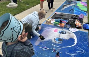 A woman draws a large animal face with chalk on the sidewalk as a child wearing a whale hat watches; colorful art supplies and another mural are nearby.