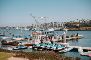 People gather on a dock with jet skis, small boats, and a fishing boat named “MAGIC” on a sunny day at a marina lined with palm trees and moored sailboats.