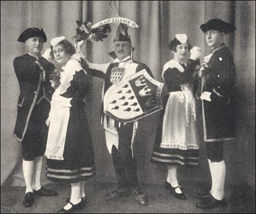 Historisches Foto von fünf Personen in historischen Kostümen mit Dreispitzhüten und Wappenschild