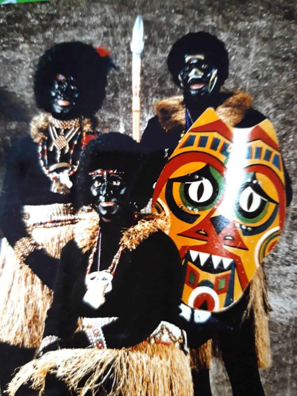 Historisches Foto von drei Personen in Kostümen mit schwarz geschminkten Gesichtern und Baströcken, dem sogenannten „Dschungelbrööder"-Kostüm, sowie einer bemalten Maske