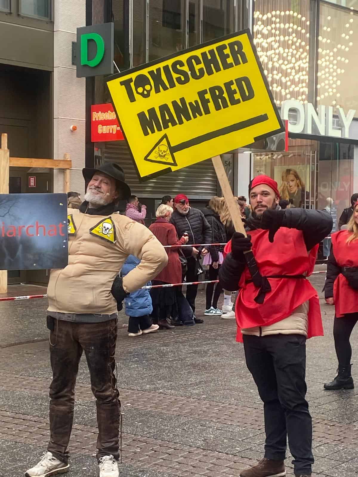 Zwei Männer bei Karnevalsveranstaltung mit Schild "Toxischer Mannfred".