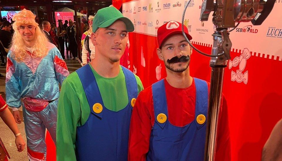Zwei Männer als Mario und Luigi verkleidet, im Hintergrund ein Mann im Retro-Trainingsanzug mit blonder Perücke