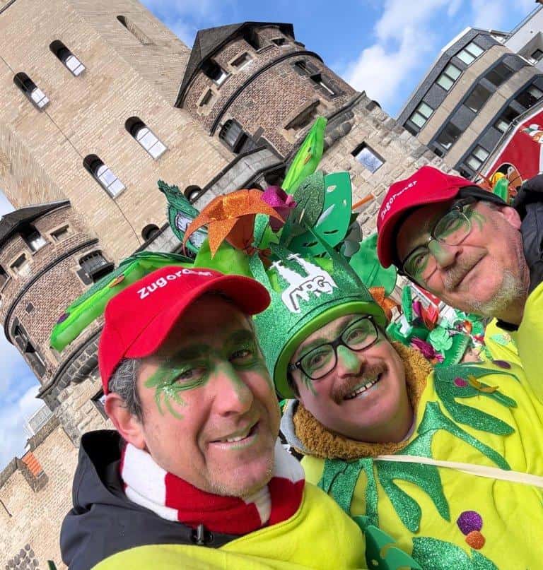 Drei Männer mit „Zugordner“-Kappen und grünen Blätterkostümen vor einem historischen Stadttor