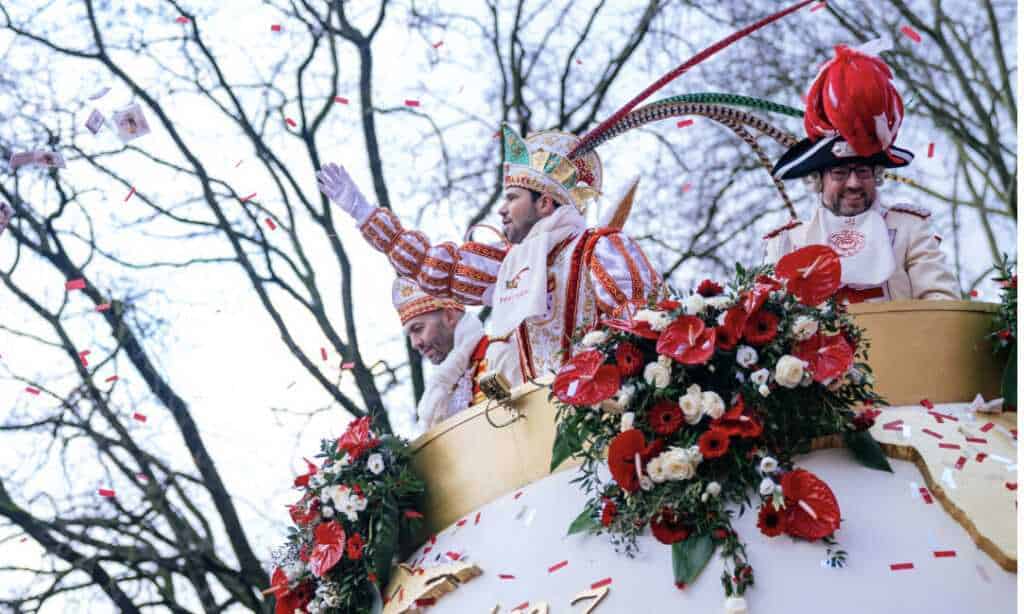 Männer in Karnevalskostümen winken von einem blumengeschmückten Festwagen, umgeben von Konfetti