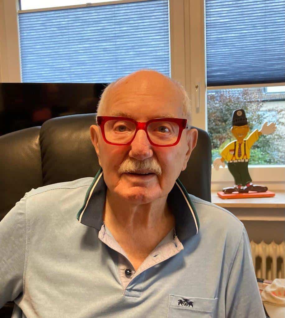 Älterer Mann mit roter Brille vor einer Webcam, im Hintergrund eine kleine Karnevalsfigur