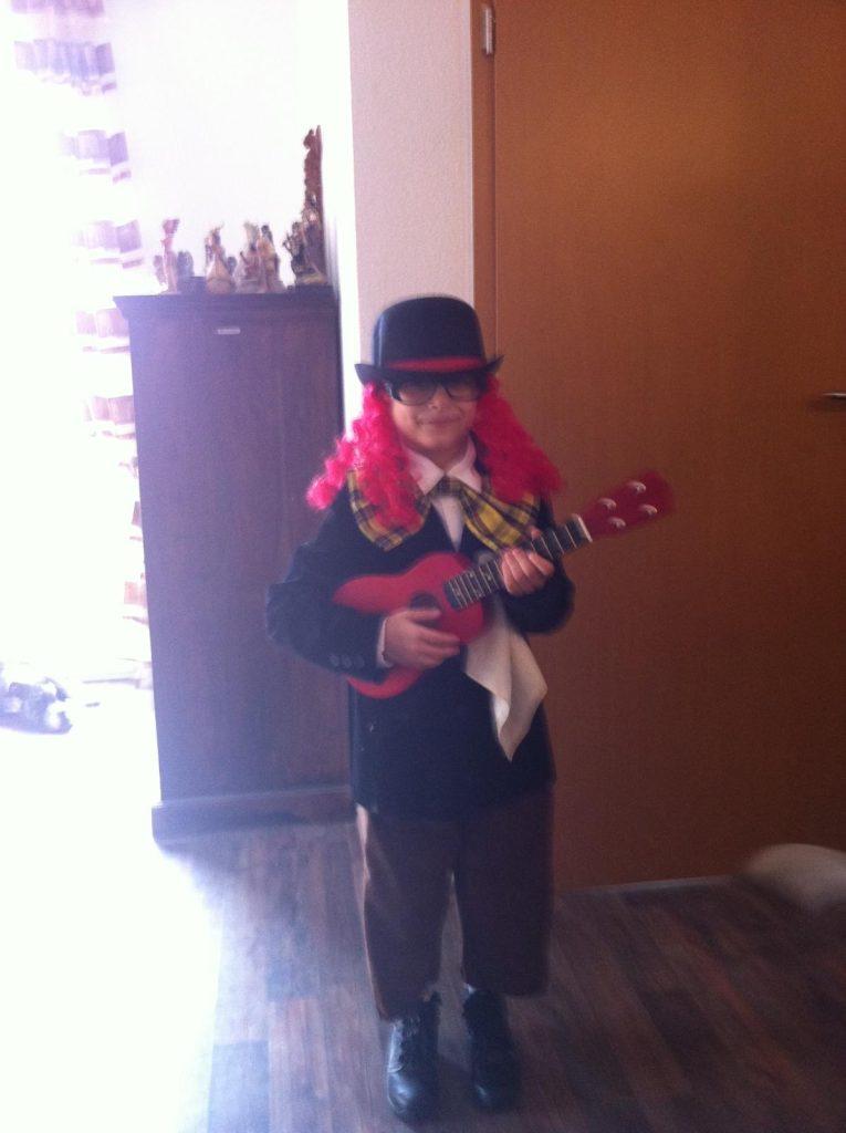 Kind im Karnevalskostüm mit Melone und roter Perücke spielt Ukulele