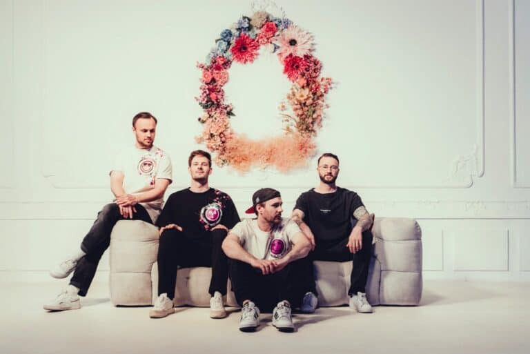 Vier Männer sitzen auf einer Couch vor einem Blumenkranz an der Wand