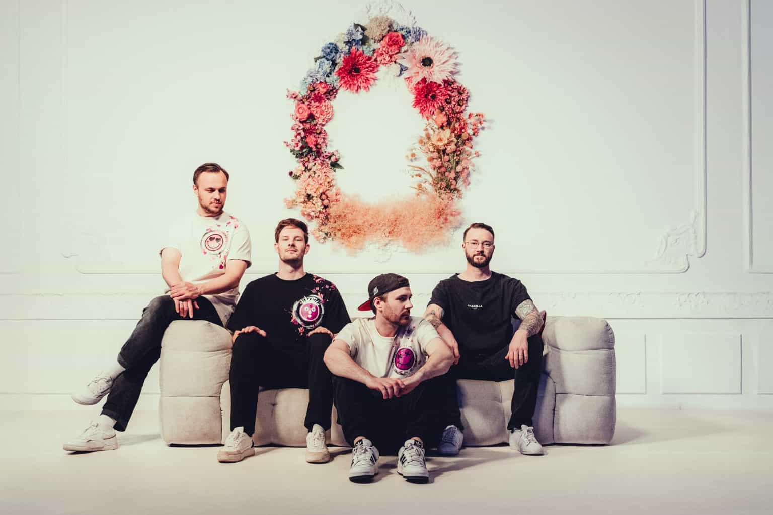 Vier Männer sitzen auf einer Couch vor einem Blumenkranz an der Wand
