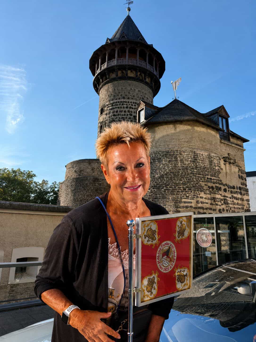 Frau hält eine Auszeichnung mit Wappen vor einem historischen Kölner Turm