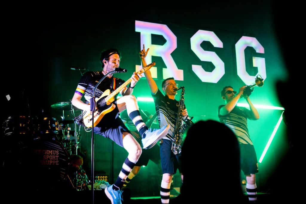 Musiker der Rhythmussportgruppe springt mit Gitarre auf der Bühne, im Hintergrund Saxophonist und Trompeter