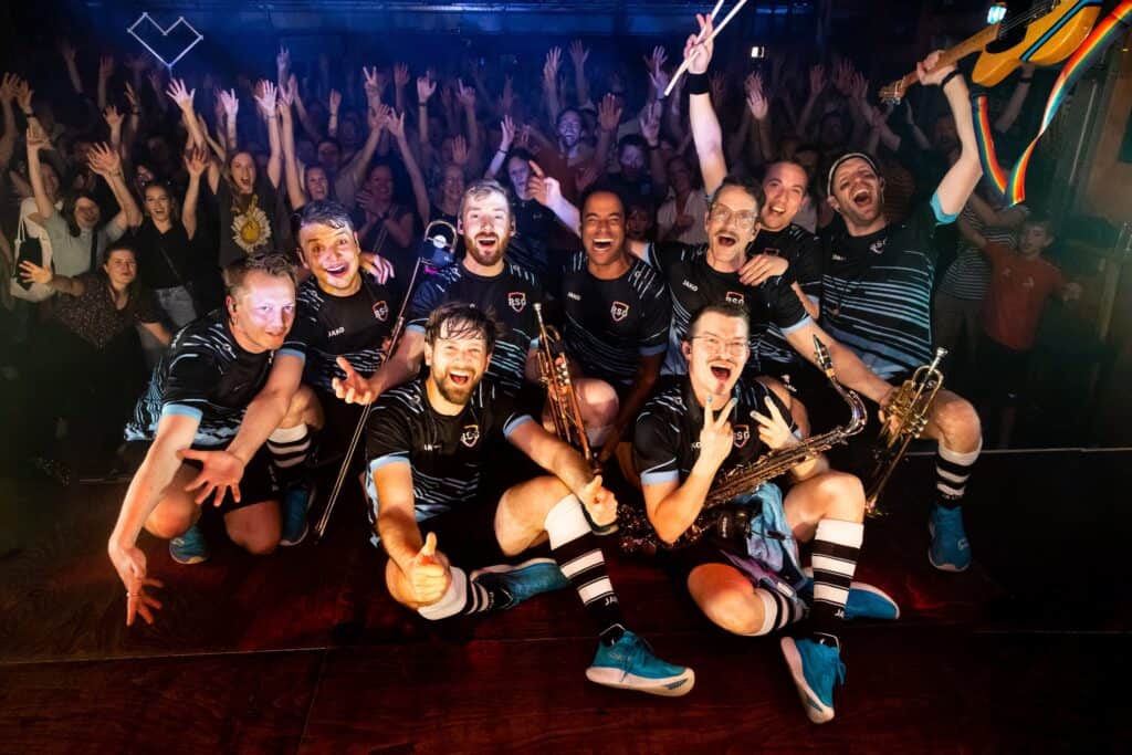 Bandmitglieder der Rhythmussportgruppe posieren mit Instrumenten vor jubelndem Publikum