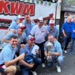 Gruppe in einheitlicher Kleidung posiert mit einem großen Pokal vor einem KWM-Banner