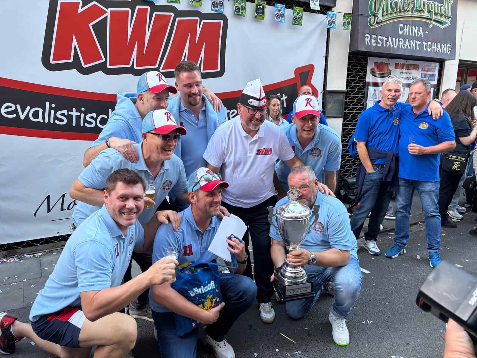 Gruppe in einheitlicher Kleidung posiert mit einem großen Pokal vor einem KWM-Banner