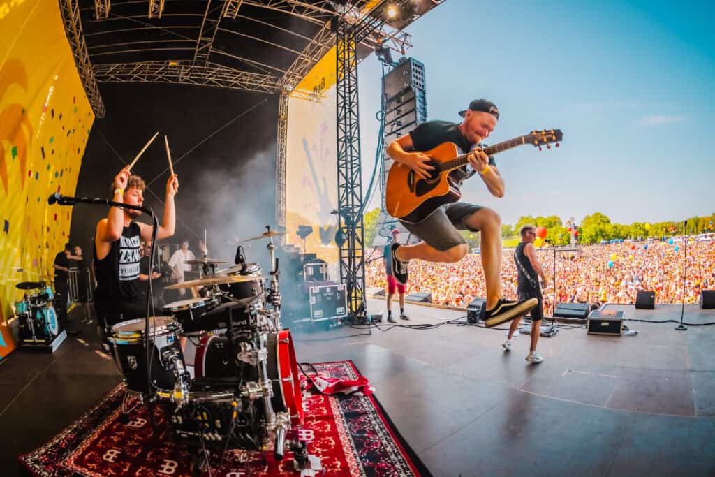 Musiker springt mit Gitarre auf einer Freiluft-Festivalbühne vor großem Publikum