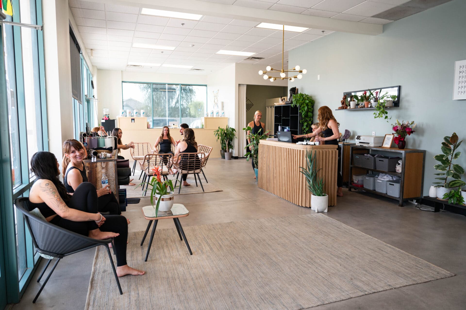 People sit and chat in a modern, brightly lit waiting area with plants, while two staff members work at a reception desk in the background.